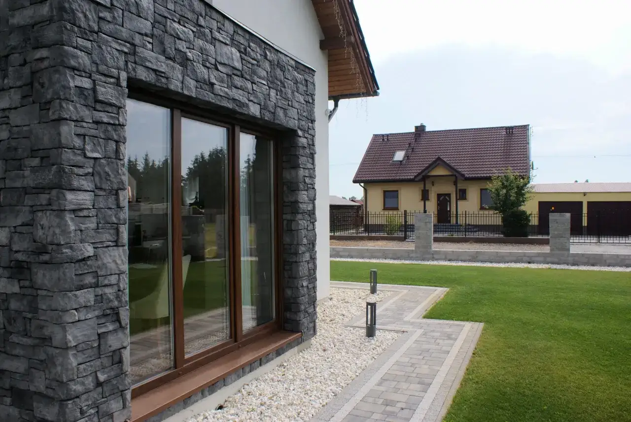 Nowoczesna elewacja z kamienia na styropianie, z dużym oknem wychodzącym na zadbany ogr&oacute;d i dom w tle.