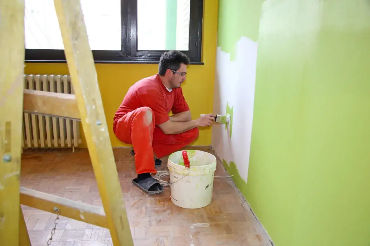 Mężczyzna w czerwonym ubraniu maluje ścianę na zielono. Zastanawiasz się, ile kosztuje malowanie pokoju?