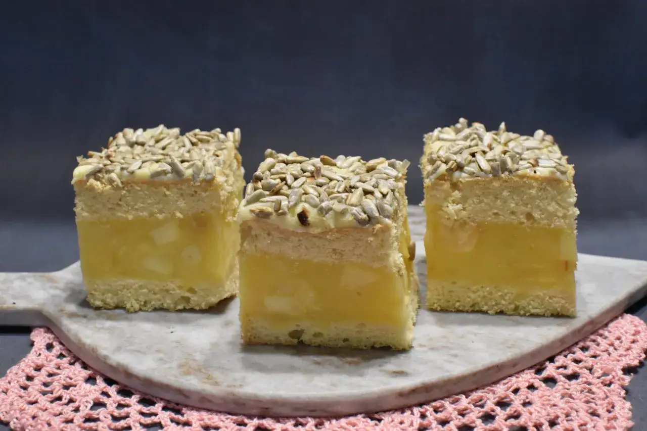 Trzy kawałki jabłecznika na biszkopcie, z kremowym nadzieniem jabłkowym i słonecznikiem na wierzchu, prezentują się apetycznie.