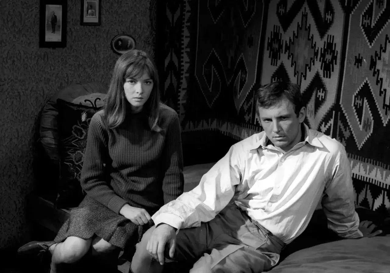 Para Eugeniusza Korczarowskiego i jego partnerkę, siedzących w ponurym wnętrzu, na tle ozdobnego gobelinu.