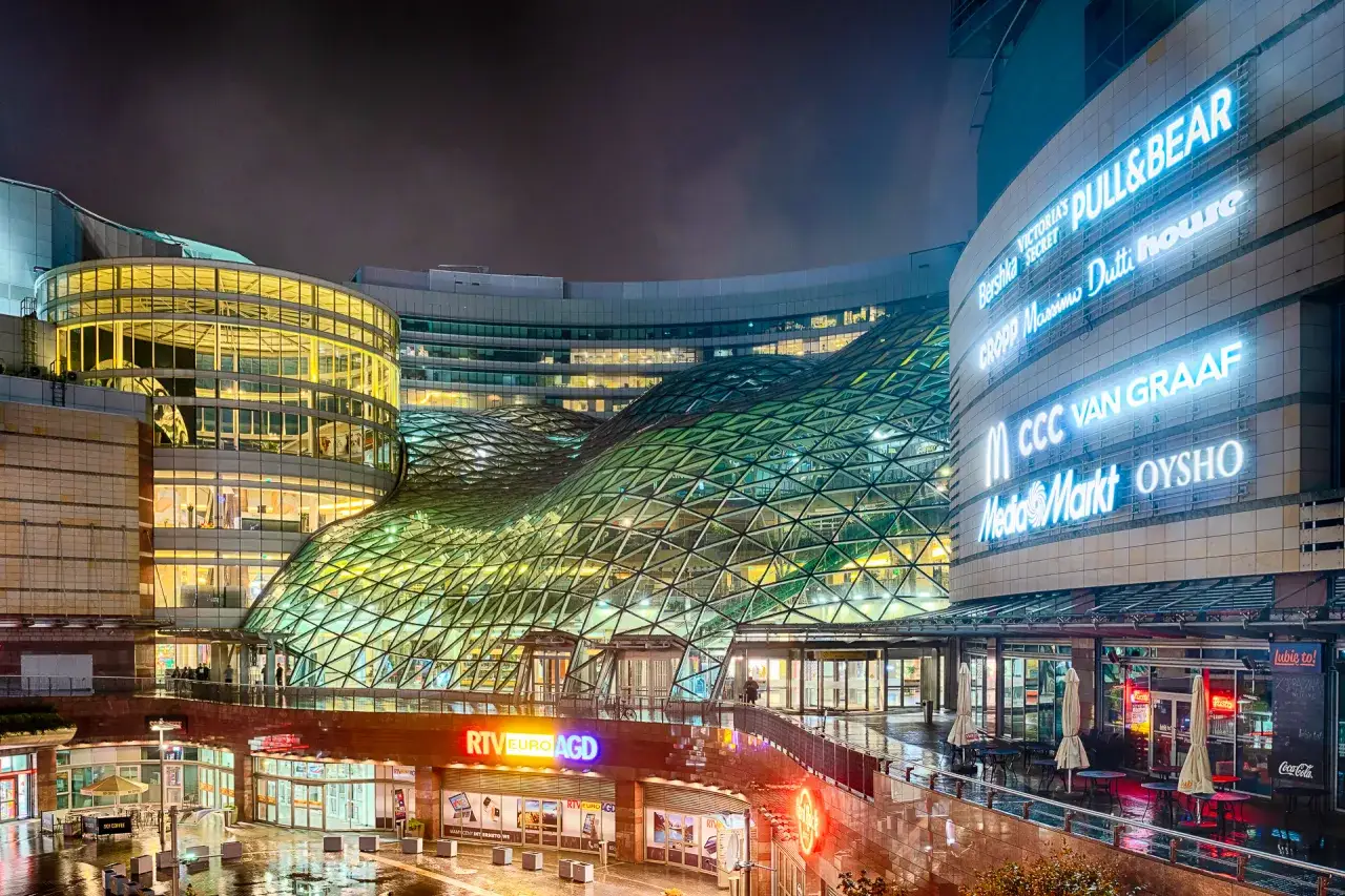 Nowoczesne centrum handlowe Złote Tarasy nocą, z charakterystyczną szklaną konstrukcją dachu i neonami sklepów.