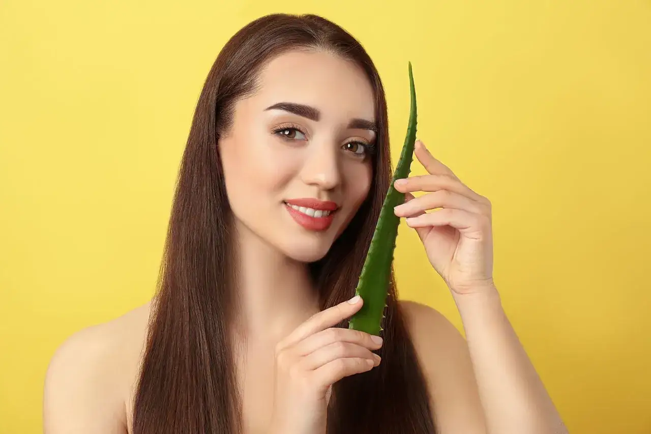 Uśmiechnięta kobieta z długimi, brązowymi włosami trzyma liść aloesu. Aloes na włosy to naturalny sposób na ich pielęgnację.