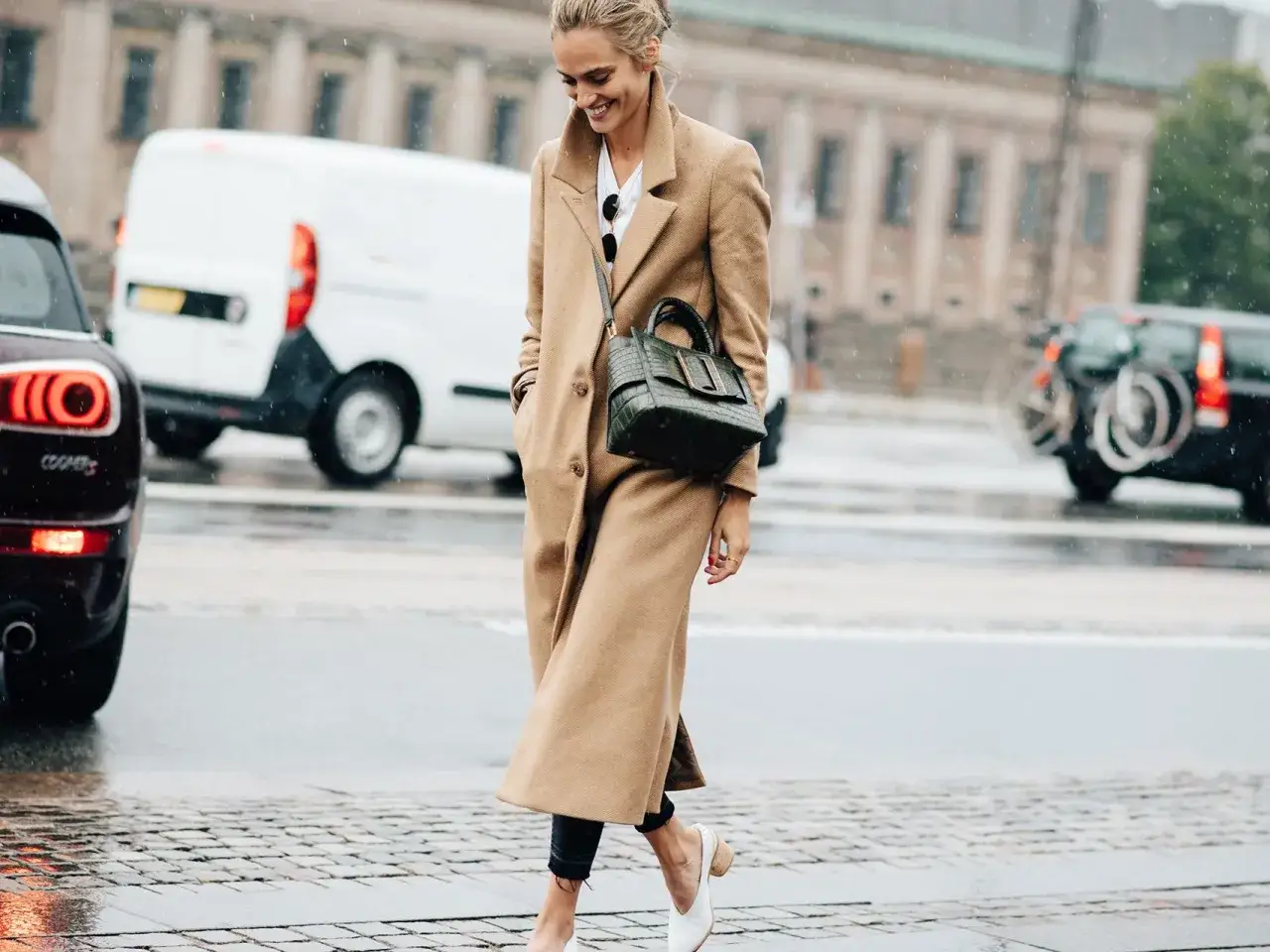Kobieta w płaszczu od skandynawskie marki odzieżowe, z zieloną torebką, idzie ulicą w deszczu.