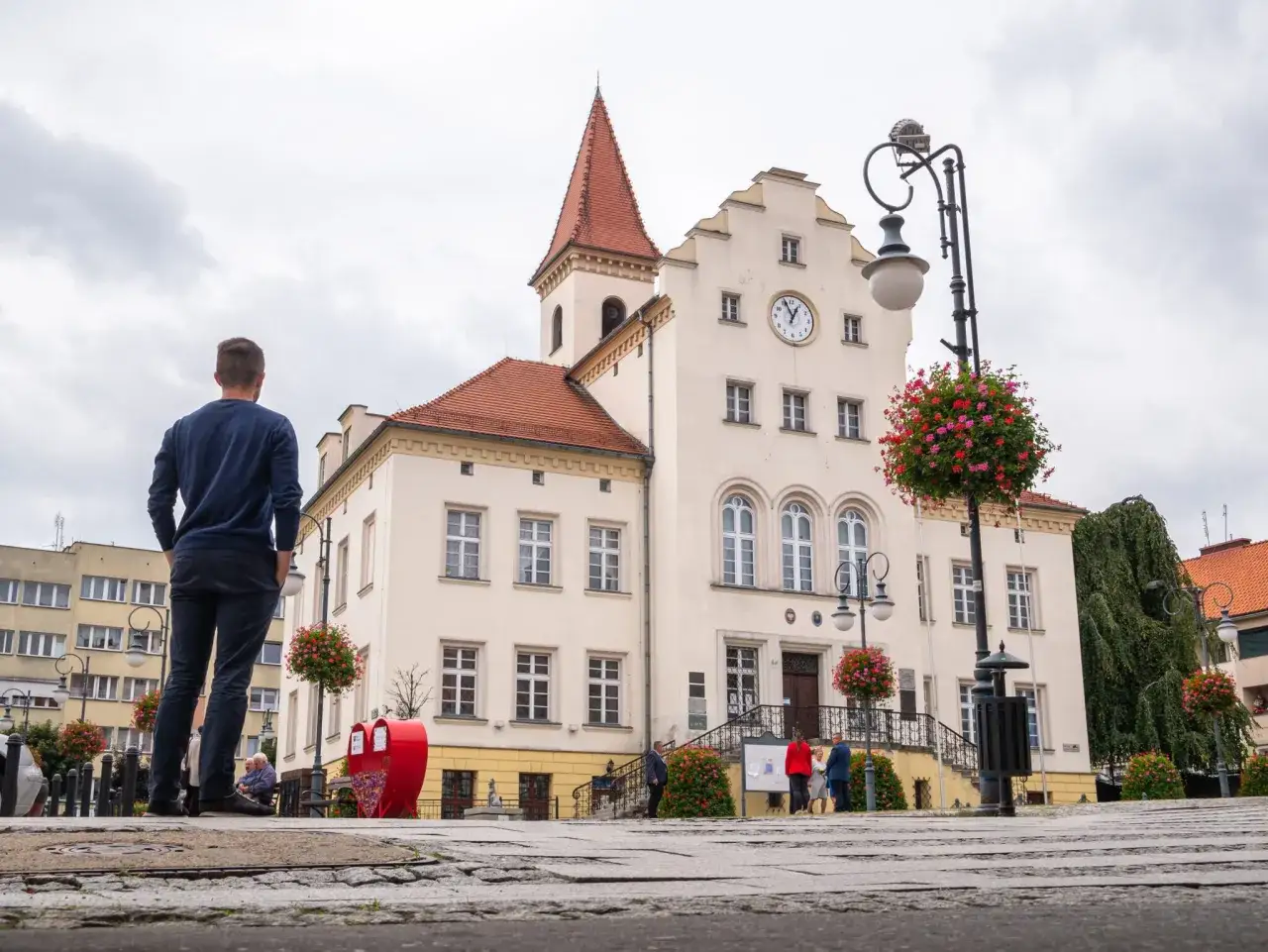 Mężczyzna patrzy na ratusz w Trzebnicy, jedno z wielu miejsc wartych zobaczenia. W tle zabytkowa architektura i kwitnące pelargonie.
