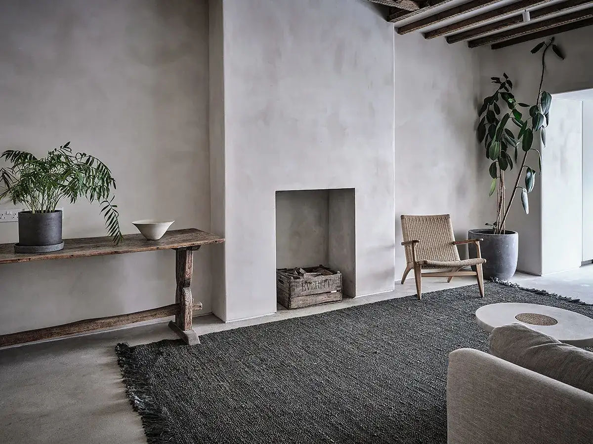 Minimalistyczny salon z betonem na ścianie, kominkiem, drewnianym stołem i plecionym fotelem.