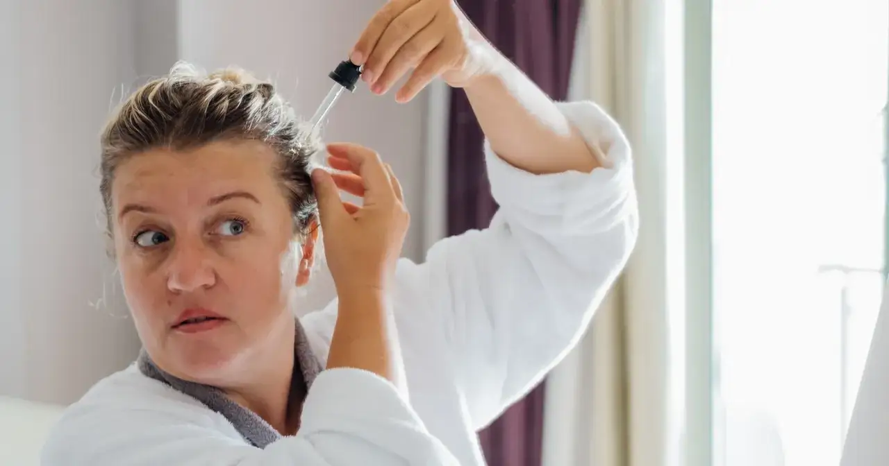 Kobieta w szlafroku aplikuje pipetką płyn na sk&oacute;rę głowy, być może eksperymentując z aspiryną na włosy dla ich wzmocnienia.