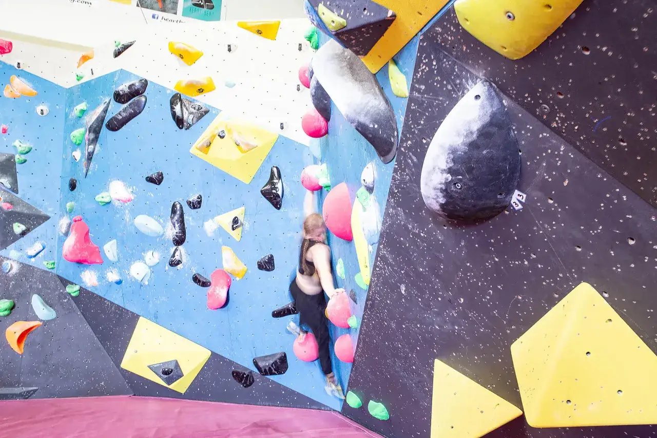 Kobieta wspina się po kolorowej ścianie wspinaczkowej. R&oacute;żne chwyty tworzą trudną trasę na tej skali boulderowej.