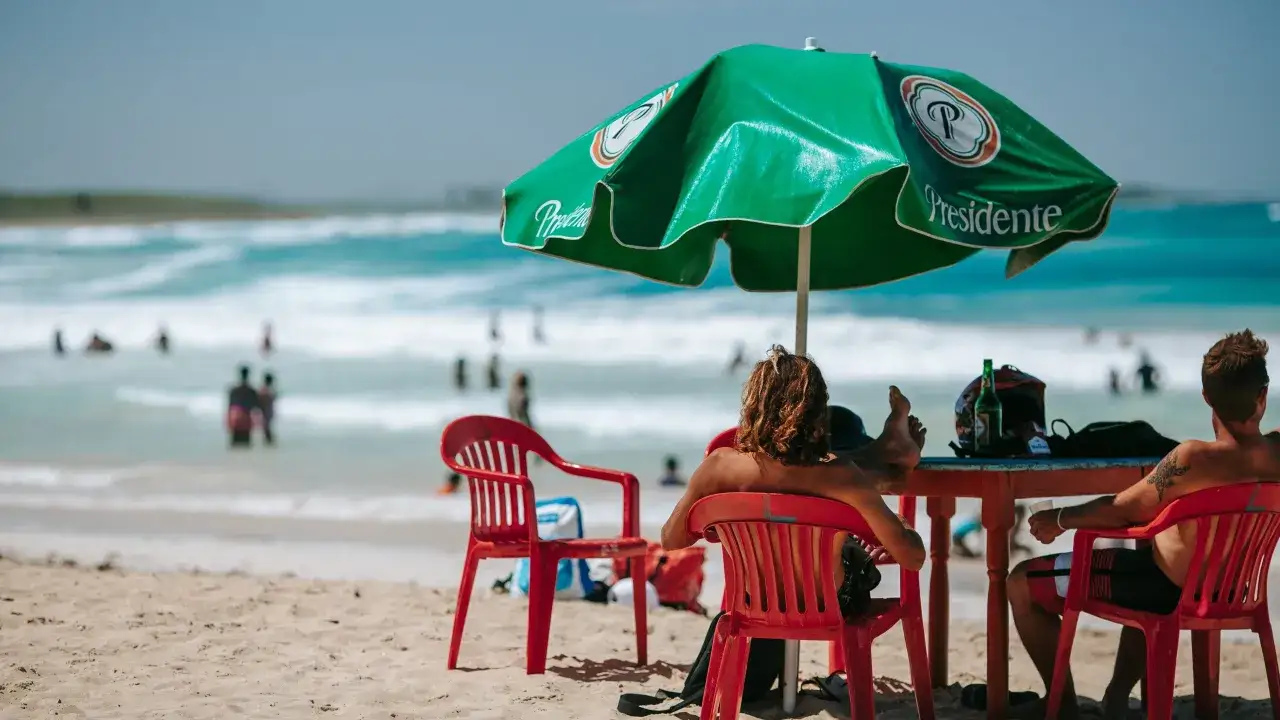 Dw&oacute;ch mężczyzn odpoczywa pod zielonym parasolem na plaży, marząc o tym, kiedy Dominikana będzie najtaniej.