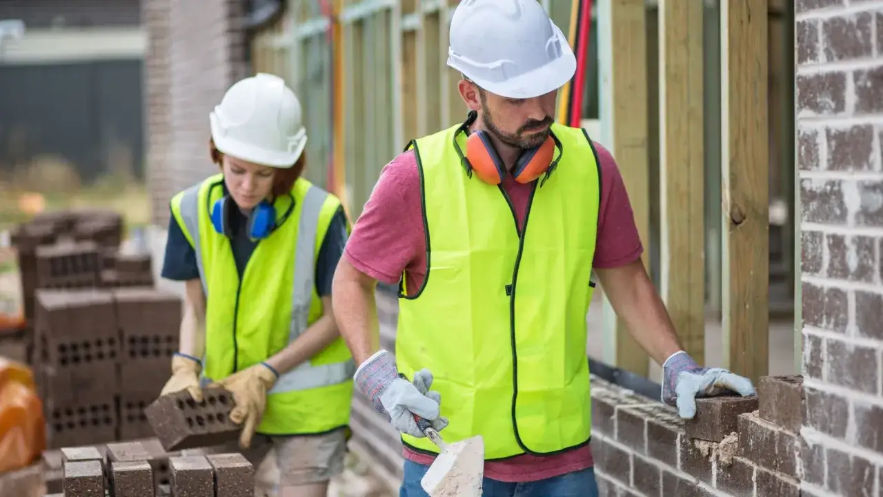 Dwóch budowlańców w kamizelkach odblaskowych układa cegły. Zastanawiasz się, ile zarabia pomocnik na budowie w Warszawie?