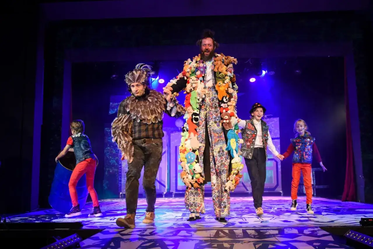 Na scenie teatru Akademia Pana Kleksa, aktorzy w barwnych strojach, w tym jeden w płaszczu z pluszaków, pozują z dziećmi.