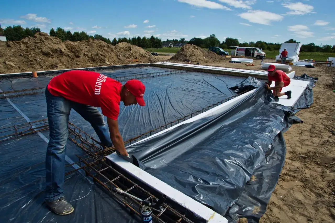 Pracownicy układają folię i styropian, przygotowując izolację płyty fundamentowej.