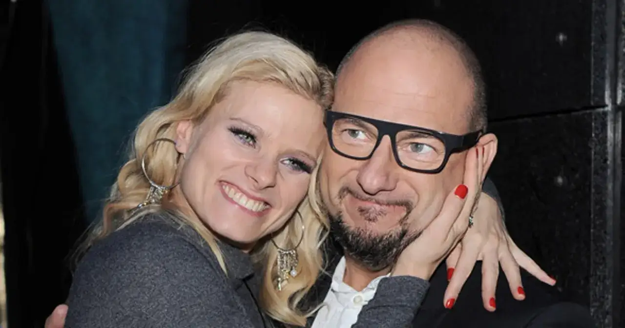 Piotr Gąsowski z partnerką, uśmiechnięci i blisko siebie. Kobieta ma blond włosy i duże kolczyki, mężczyzna okulary.