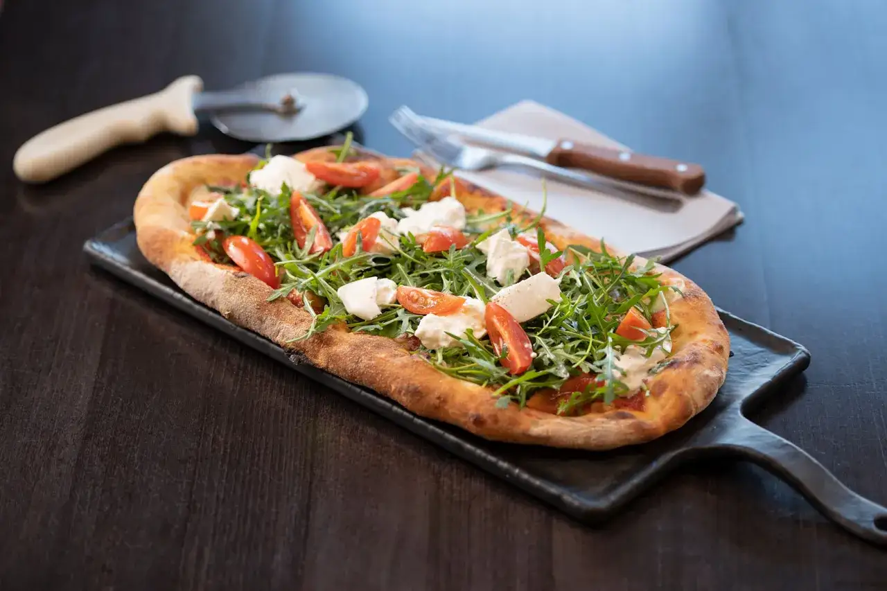 Pyszny ser do pizzy z rukolą i pomidorkami na czarnej desce. Obok leży n&oacute;ż i widelec.