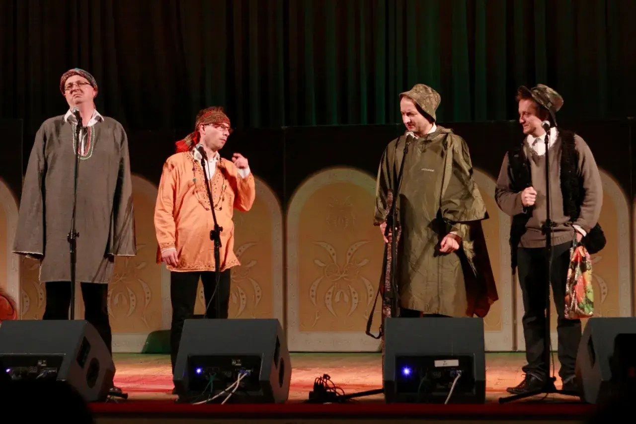 Czterech mężczyzn na scenie, w tym jeden w pomarańczowej tunice, jakby z kabaretu moralnego niepokoju.