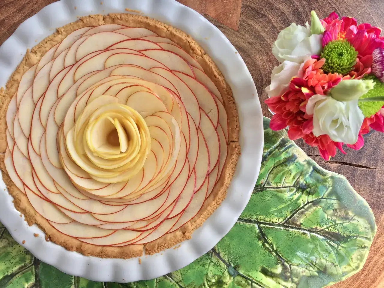 Przepis na kruche ciasto z jabłkami ułożonymi w kształt róży. Idealne na deser, obok bukiet kolorowych kwiatów.