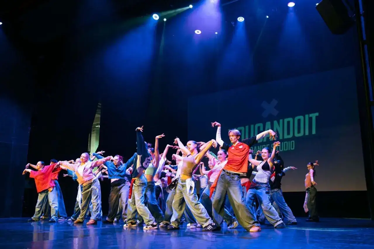 Grupa młodych tancerzy na scenie, wykonująca energiczny układ. Choreograf stworzył dynamiczną kompozycję ruch&oacute;w.