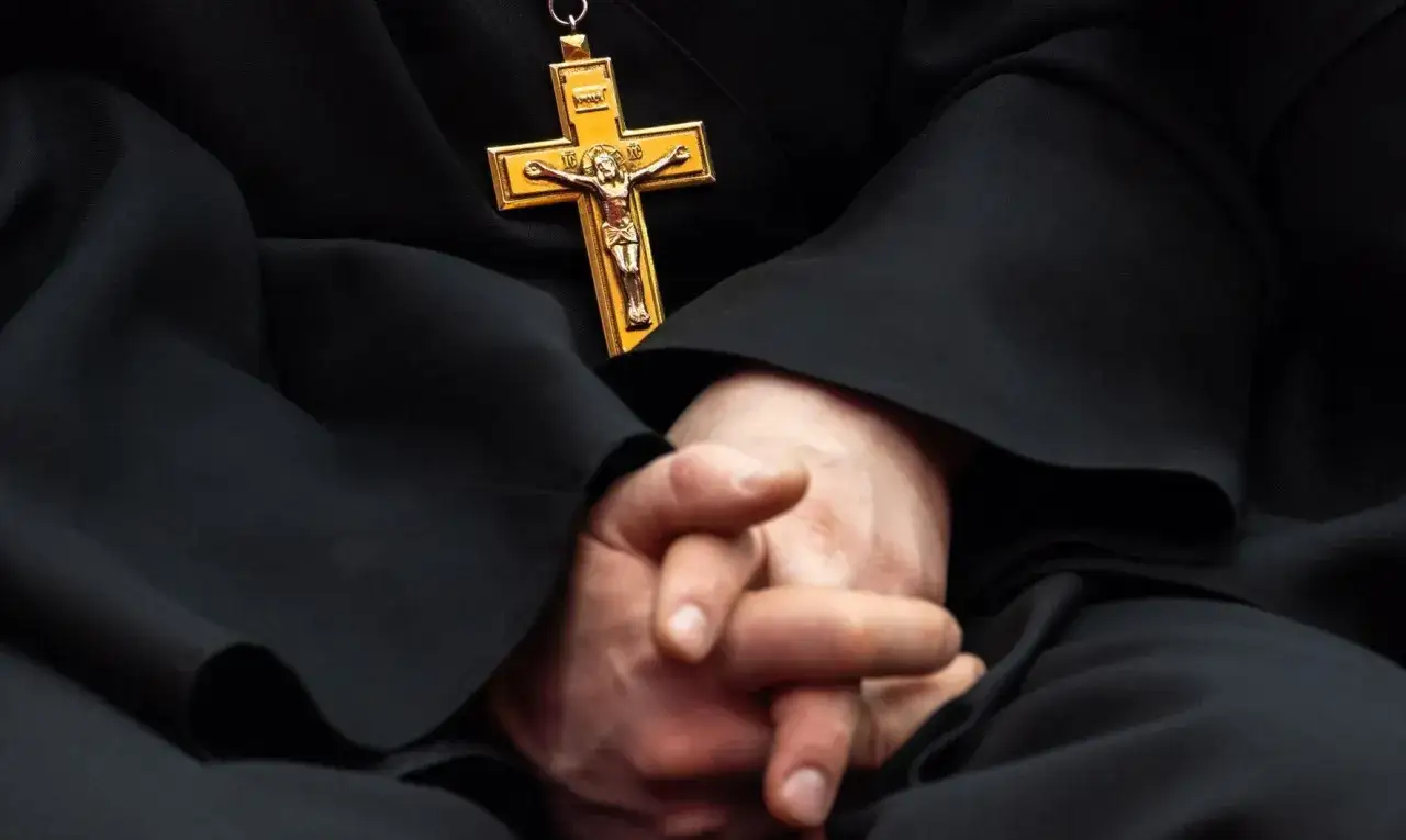 Złożone dłonie duchownego z krzyżem na piersi. Wyniki badania CBOS dotyczące praktyk religijnych w Polsce.