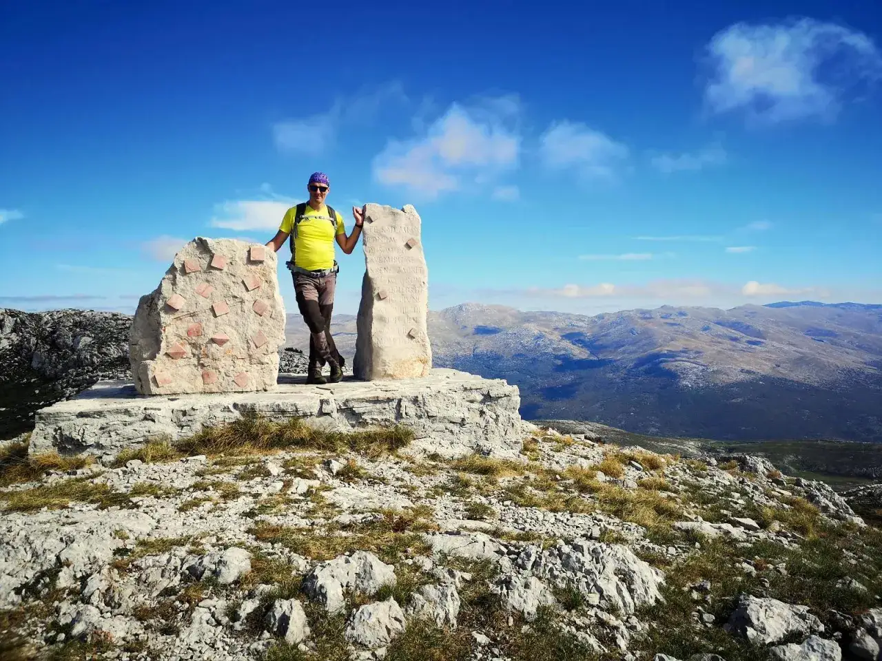 Mężczyzna w żółtej koszulce stoi na szczycie góry, opierając się o kamienne monumenty. W tle rozciąga się malowniczy krajobraz górski.