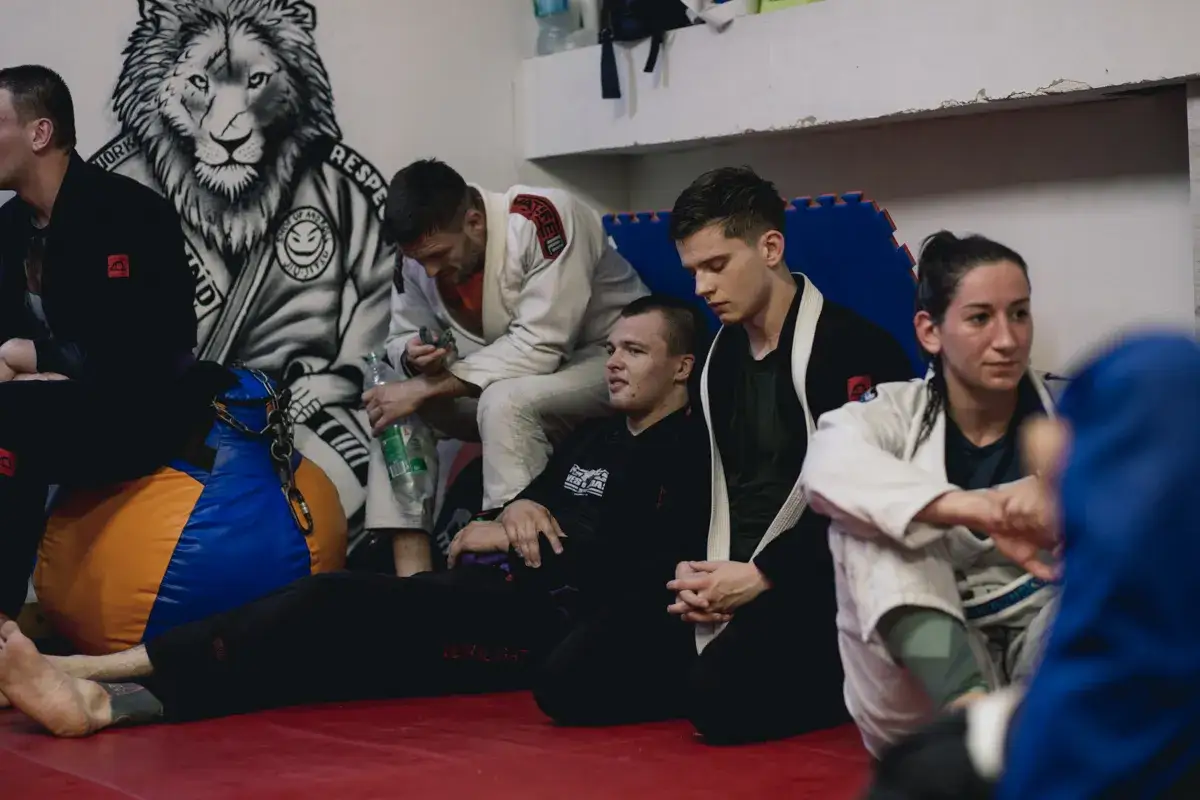 Grupa os&oacute;b w strojach do jiu jitsu odpoczywa na macie. W tle mural z lwem.