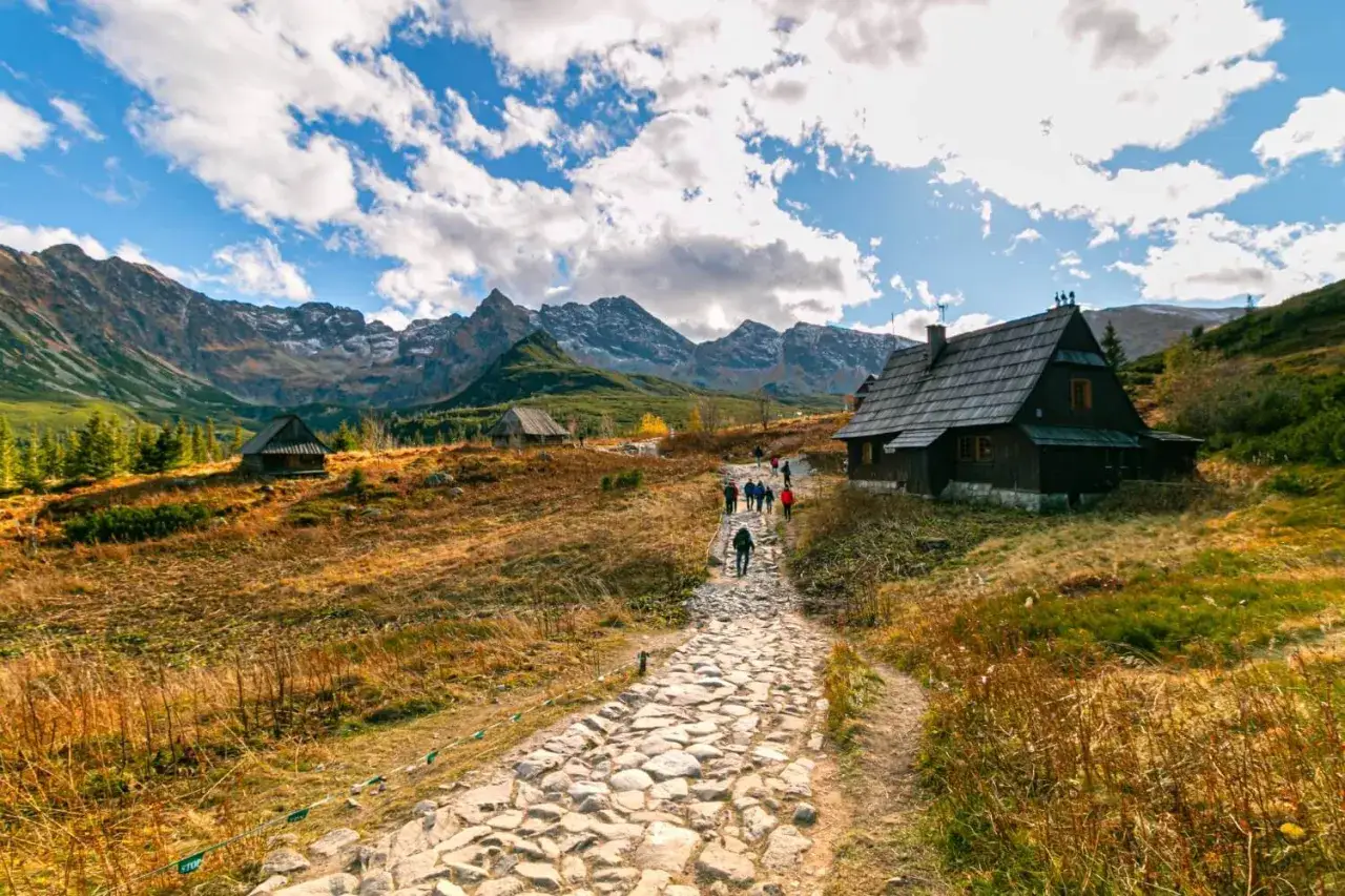 Kamienna ścieżka prowadzi do tradycyjnych **hal w Tatrach**. W oddali widać g&oacute;ry, a po bokach kamienne chaty i turyst&oacute;w.