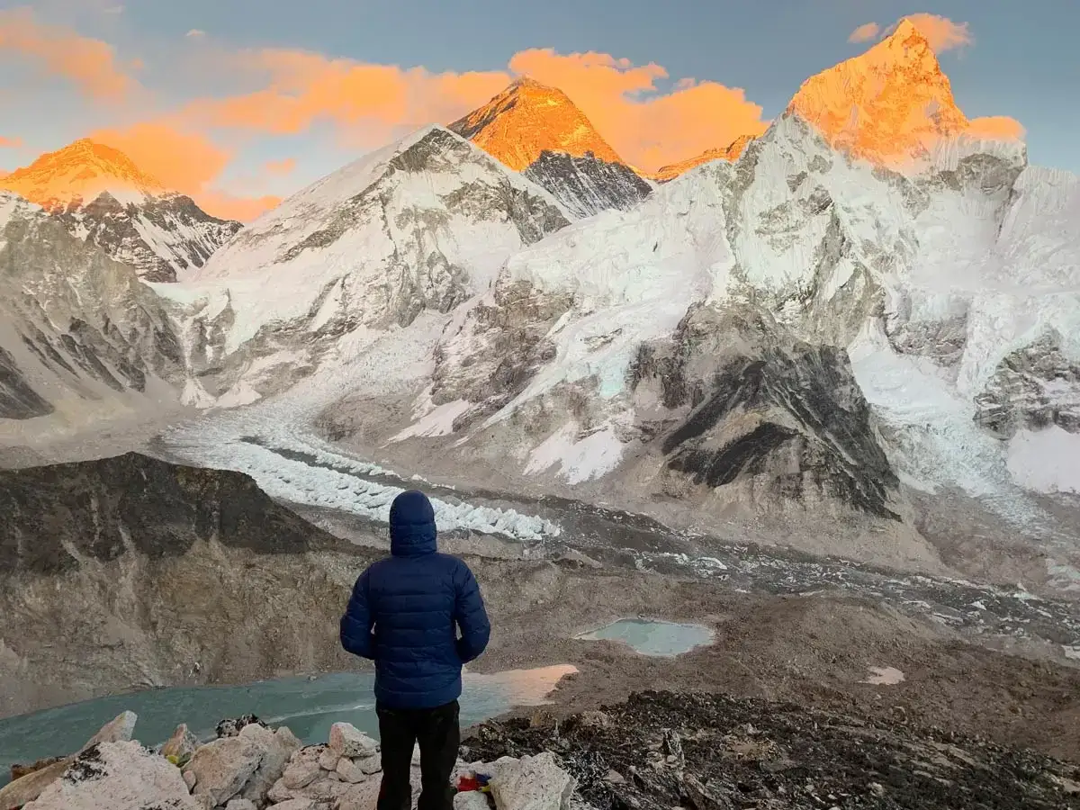 Osoba w niebieskiej kurtce podziwia ośnieżone szczyty Himalaj&oacute;w o wschodzie słońca. Mount Everest, w jakim kraju jest ten majestat, znajduje się na granicy Nepalu i Chin.
