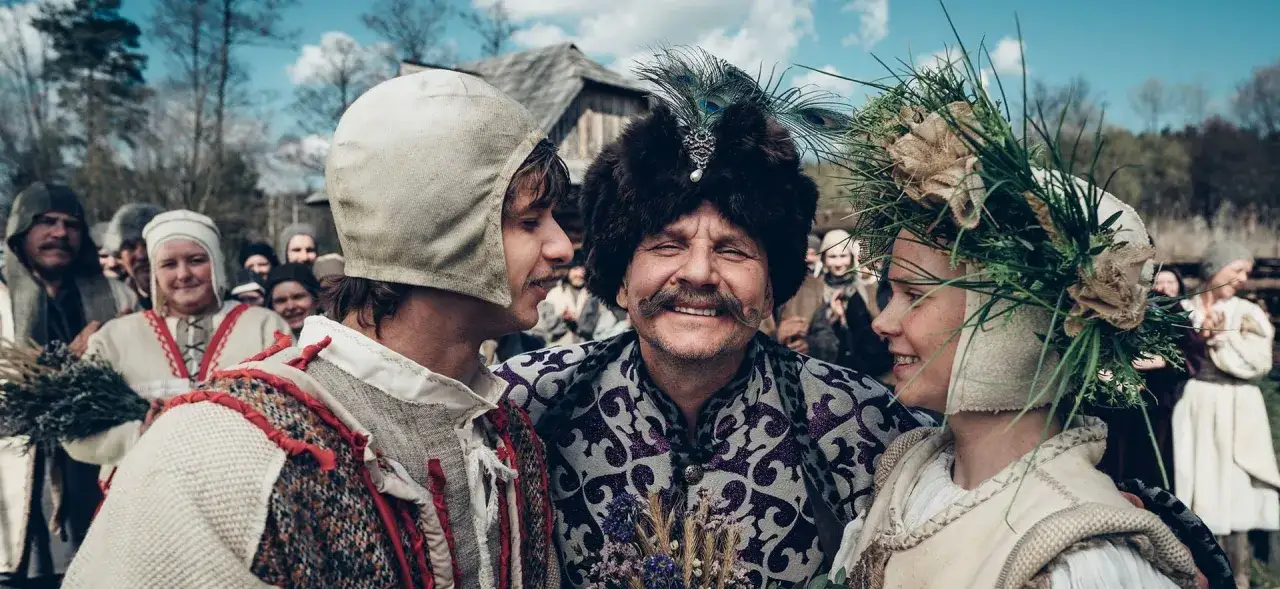 Trzech ludzi w strojach ludowych, uśmiechniętych, na tle tłumu. Scena jak z filmu o historii Polski, stand up.