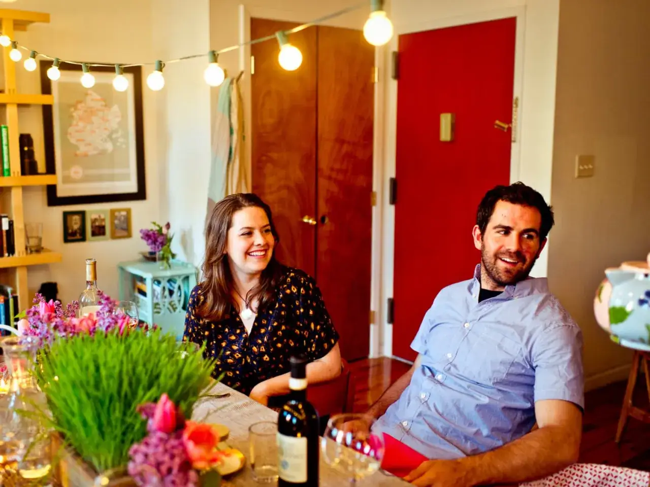 Uśmiechnięci ludzie przy stole, przygotowani na efektowna kolacja dla znajomych. W tle czerwone drzwi i girlanda z żarówek.