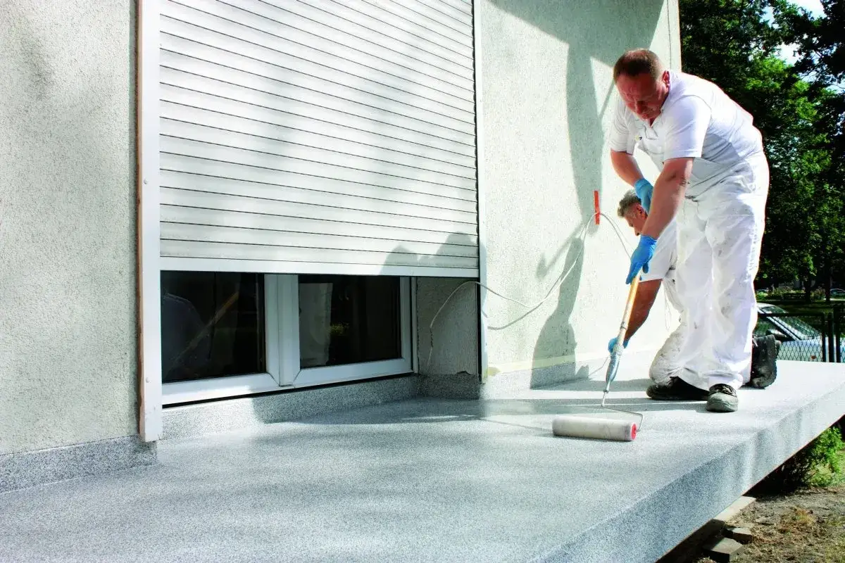 Dw&oacute;ch mężczyzn w białych kombinezonach maluje balkon. Na pierwszym planie widać wałek do malowania, a na ścianie okno z roletą. Powierzchnia balkonu pokryta jest gumolitem.