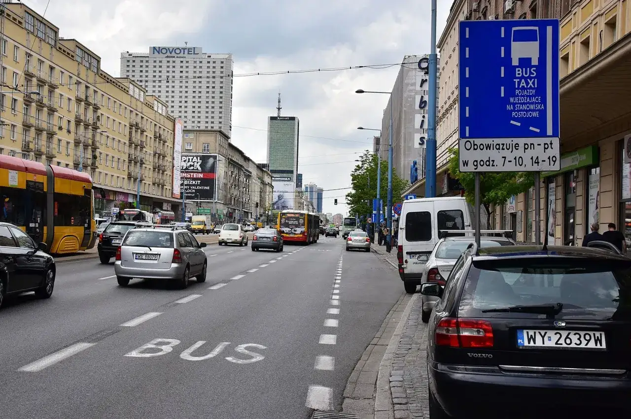 Znak informuje, kto może jeździć buspasem: autobusy i taksówki w określonych godzinach. Na drodze ruch uliczny.
