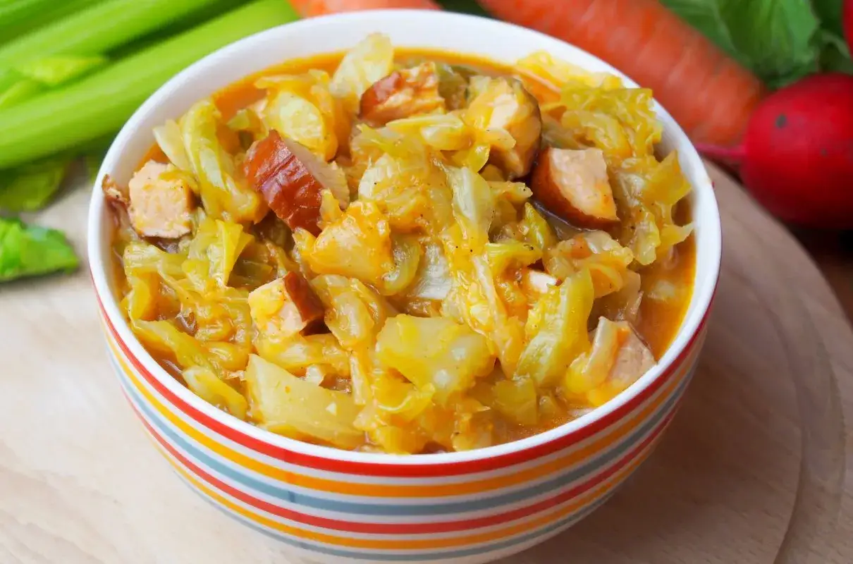 Miska pełna bigosu staropolskiego z kawałkami kiełbasy i kapusty. Obok marchew, rzodkiewka i łodygi selera.