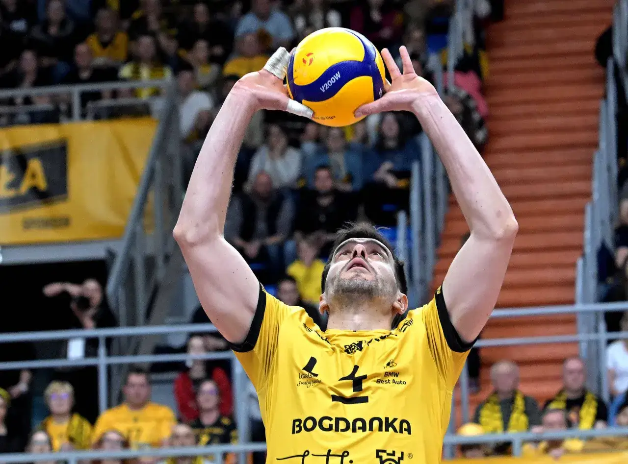 Siatkarz w żółtej koszulce z numerem 4, przygotowuje się do zagrywki. Kibice na trybunach, emocje związane z Plus Ligą i transferami.