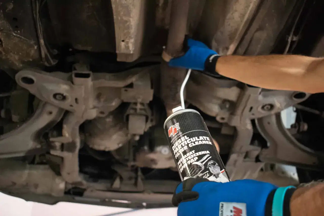 Ręce w niebieskich rękawiczkach aplikują preparat do czyszczenia DPF pod samochodem.