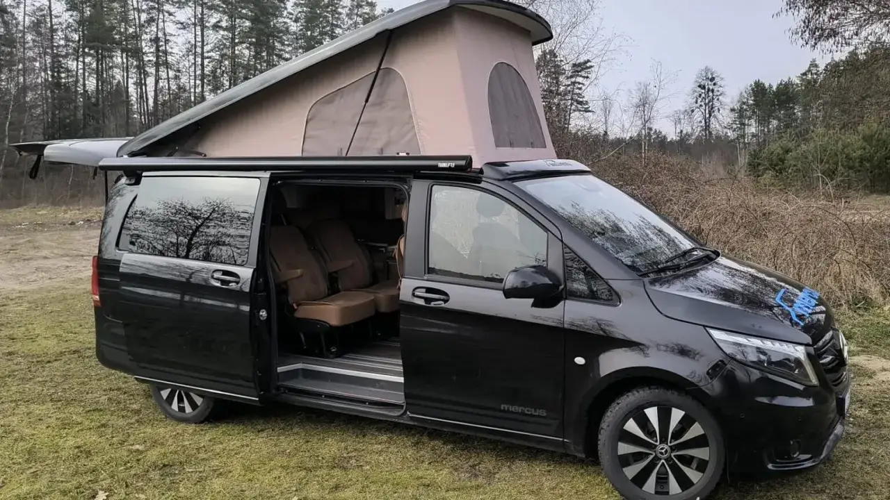 Czarny camper dwuosobowy z podnoszonym dachem, gotowy na przygodę w lesie. Wygodne fotele i przestronne wnętrze zapraszają do podróży.