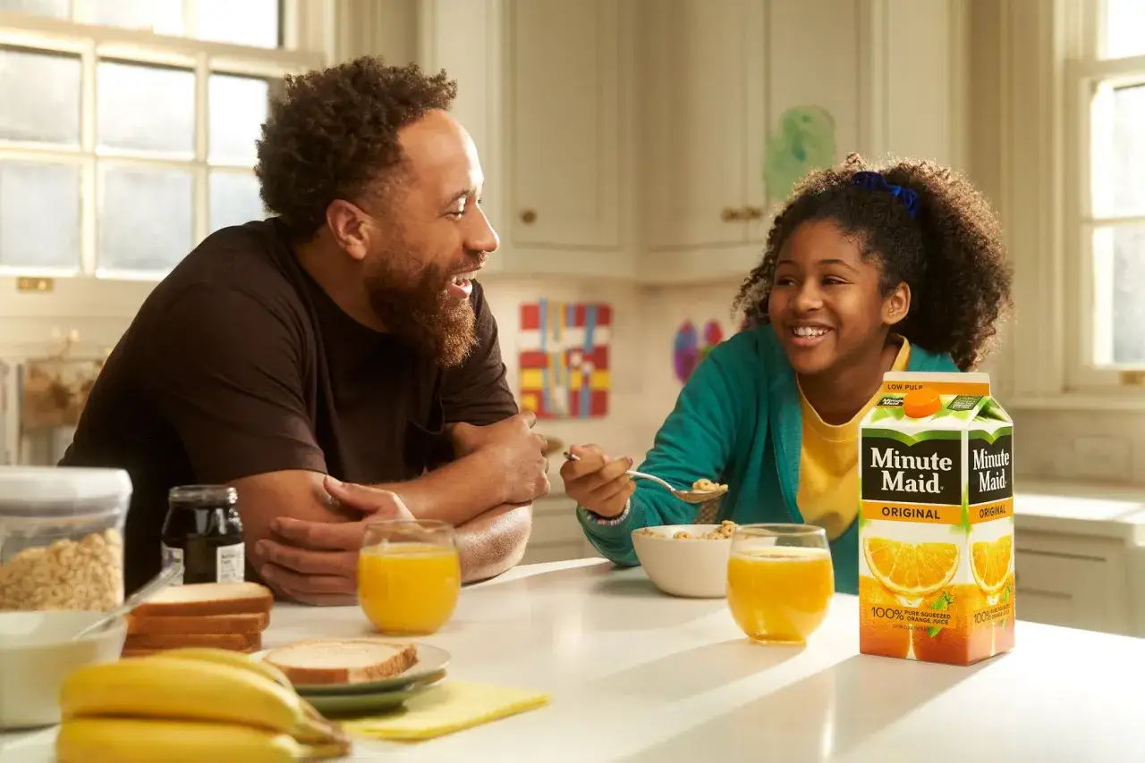 Radosna fotografia reklamowa: ojciec i c&oacute;rka jedzą śniadanie, popijając sok pomarańczowy Minute Maid.
