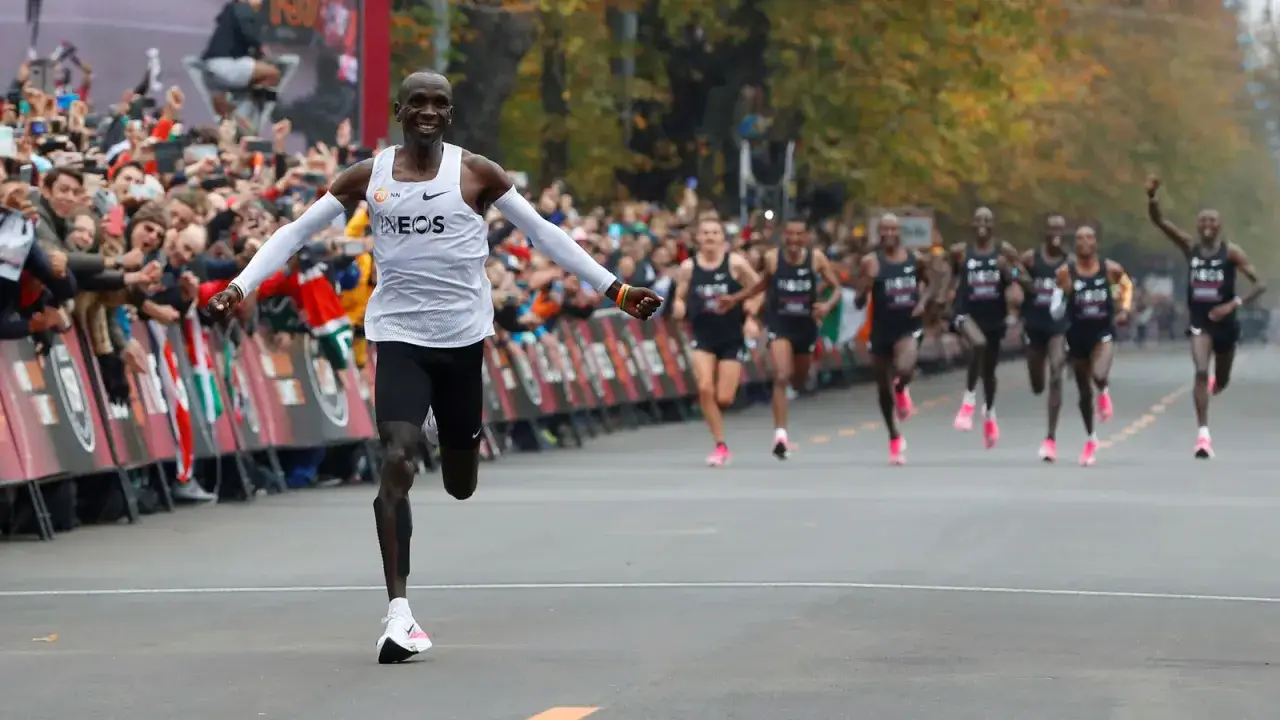 Eliud Kipchoge biegnie do mety, świętując swój **maraton rekord świata**. Tłum kibiców dopinguje go zza barierek.