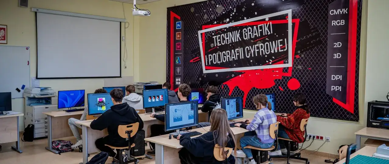 Nowoczesna klasa szkolna z uczniami pracującymi przy komputerach. Na ścianie baner