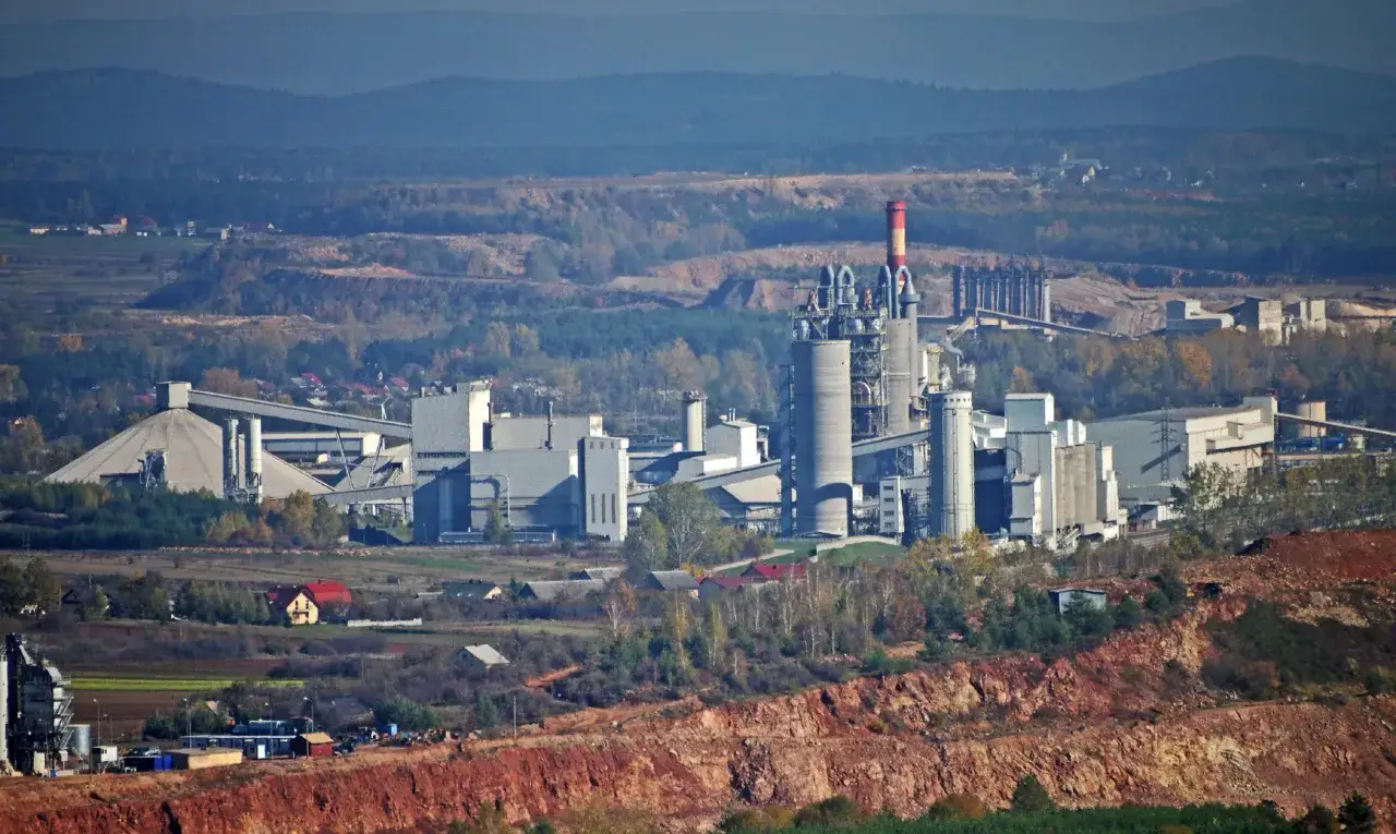 Krajobraz przemysłowy z cementowniami w Polsce, z charakterystycznymi silosami i kominami, na tle pagórków i lasów.