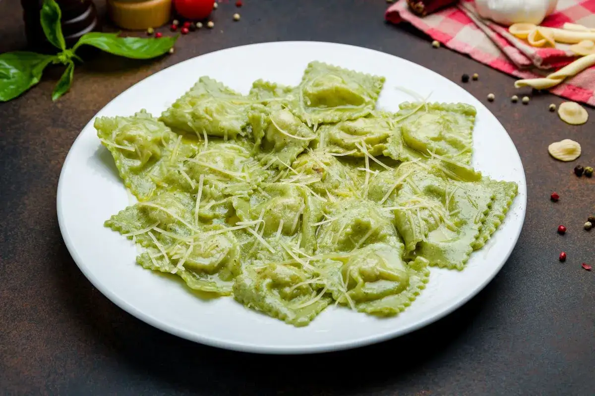 Zielone ravioli z ricottą, polane kremowym sosem i posypane startym parmezanem, prezentują się apetycznie na białym talerzu.