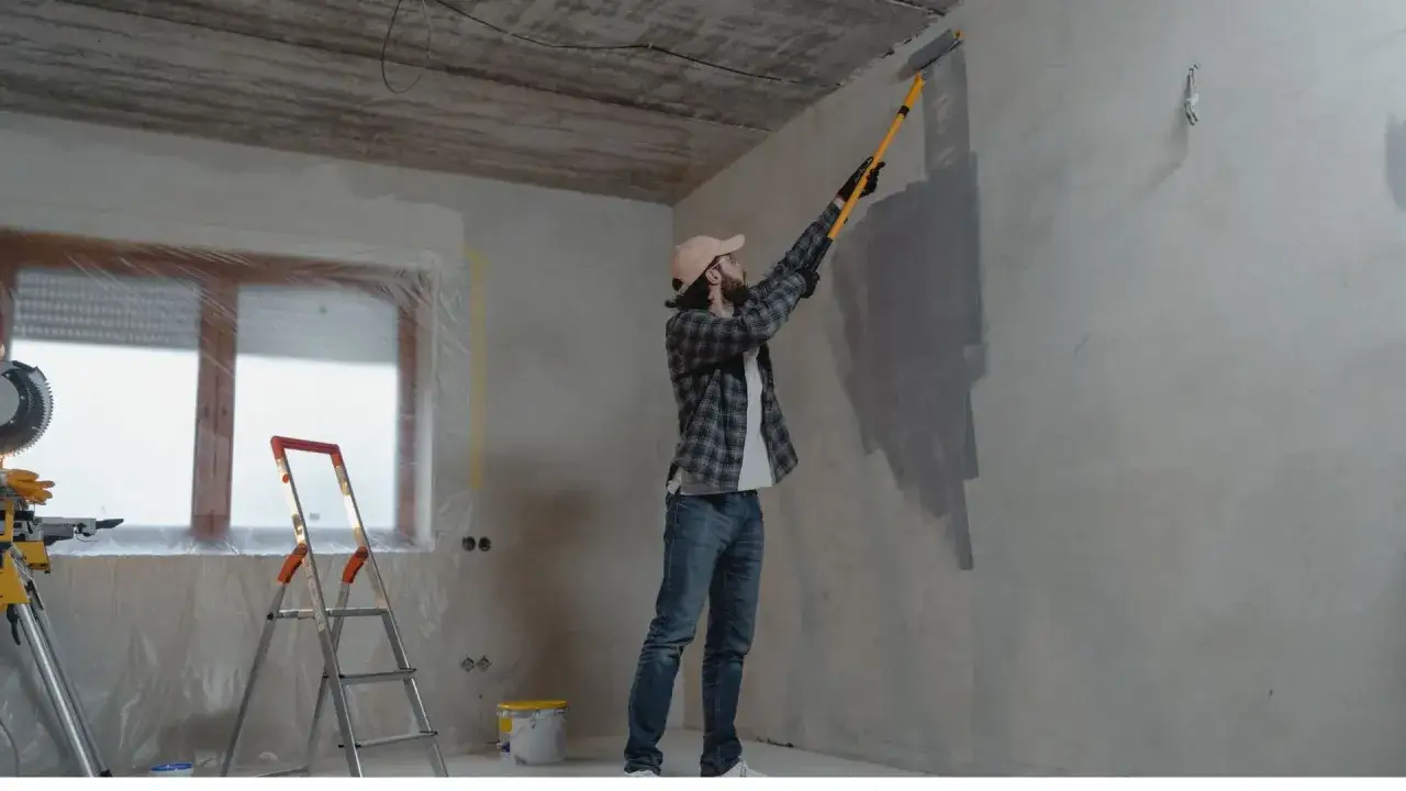 Mężczyzna maluje ścianę wałkiem. Obok drabina i wiadro z farbą. Widać, ile farby potrzeba na m2.
