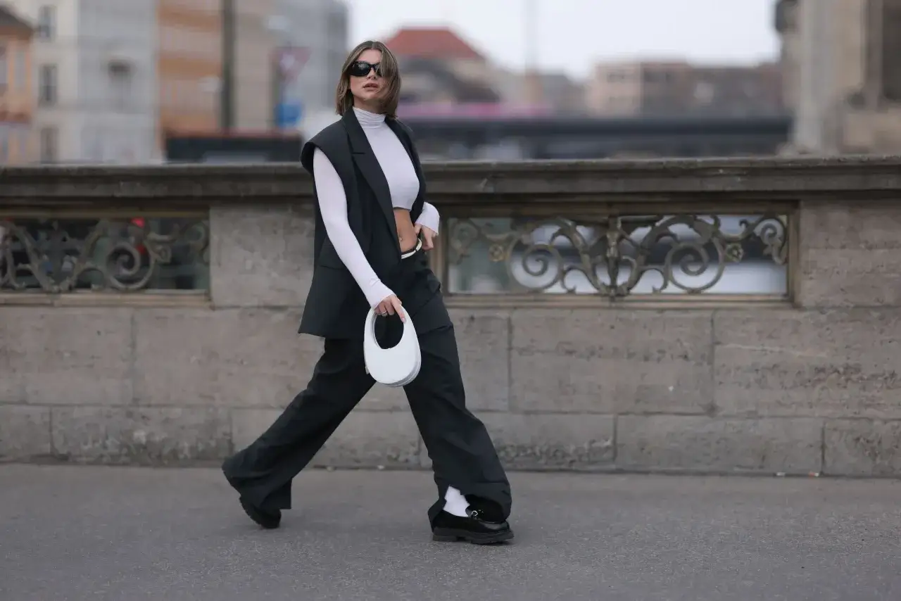 Kobieta w czarnym garniturze i białym golfie, z białą torebką, prezentuje stylową propozycję: jakie buty do szerokich spodni? Cz&oacute;łenka na grubej podeszwie i białe skarpetki.