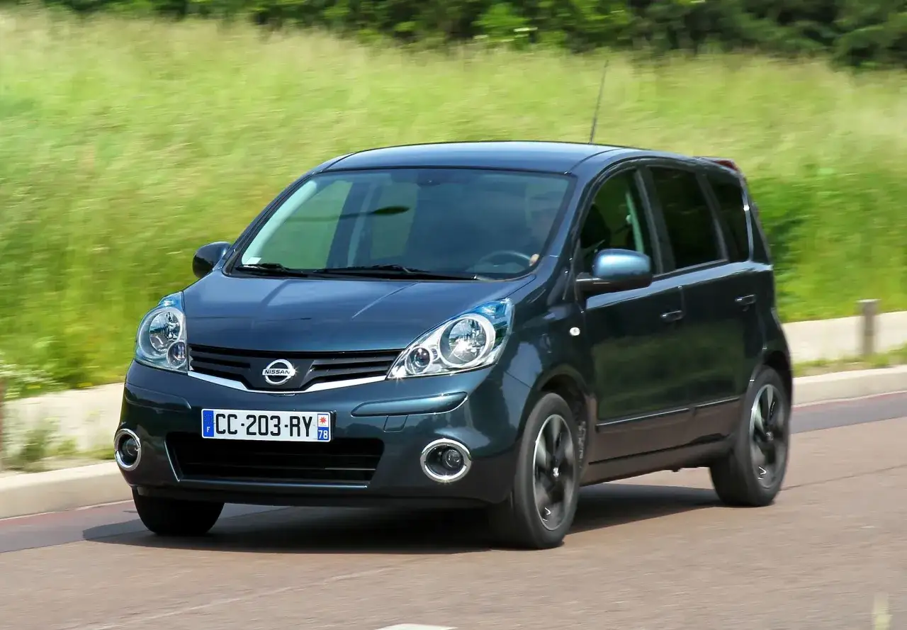 Ciemnozielony Nissan Note jedzie po drodze, otoczony zielenią.