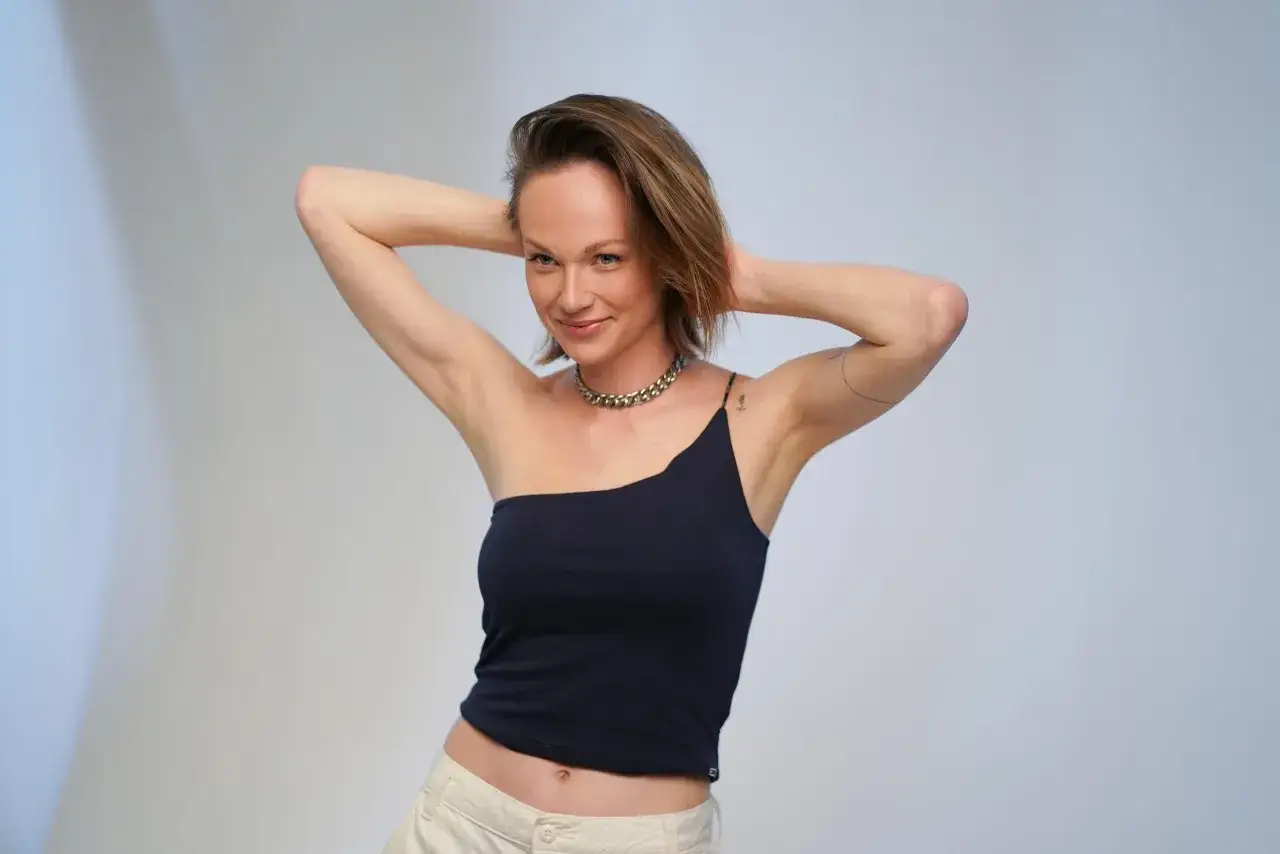 Aleksandra Adamska w czarnym topie na jedno ramię i beżowych spodniach, z rękami za głową, uśmiecha się do obiektywu.