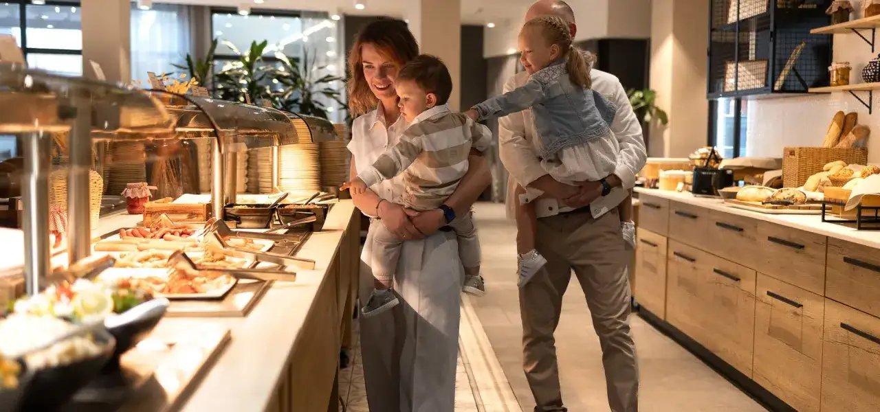 Rodzina wybiera jedzenie z bufetu. Dzieci na rękach rodzic&oacute;w, obok bogaty wyb&oacute;r potraw. To prawdziwy **ultra all inclusive**!