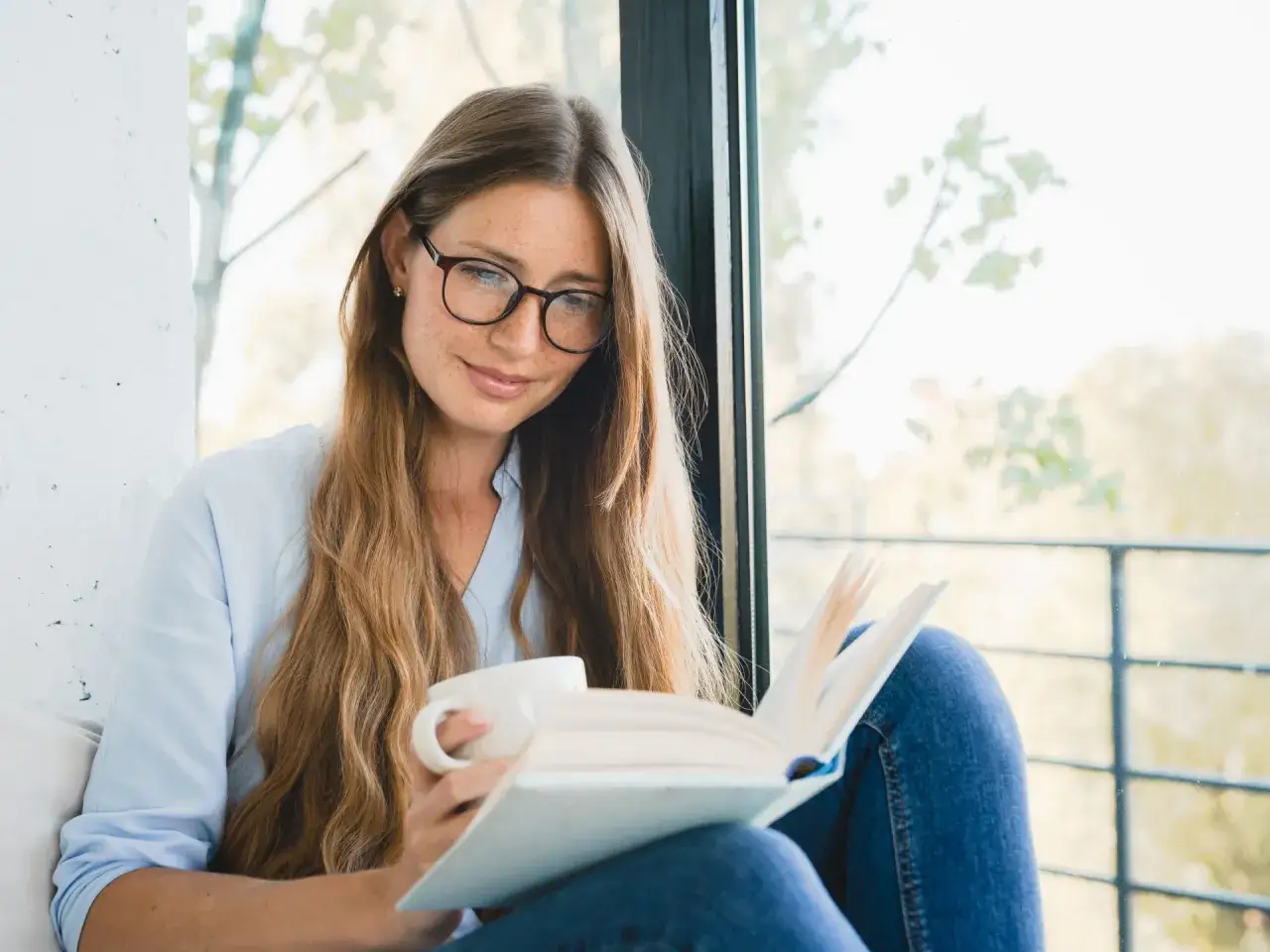 Kobieta w okularach, z kubkiem kawy, czyta książkę. Jest prawdziwym fanem dobrej literatury.