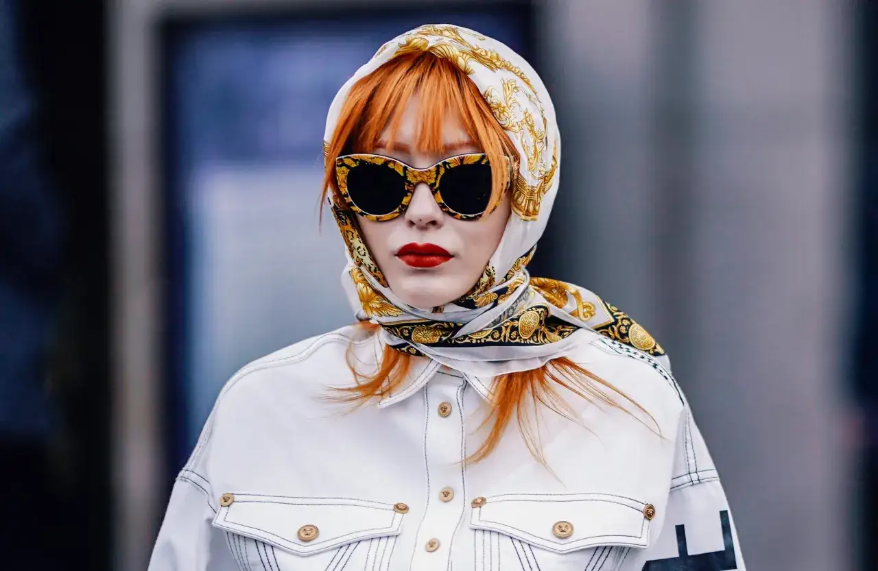 Elegancka kobieta z rudymi włosami, w okularach i chustce na głowie. Pokazuje, jak zawiązać chustę na głowie, tworząc stylowy look.