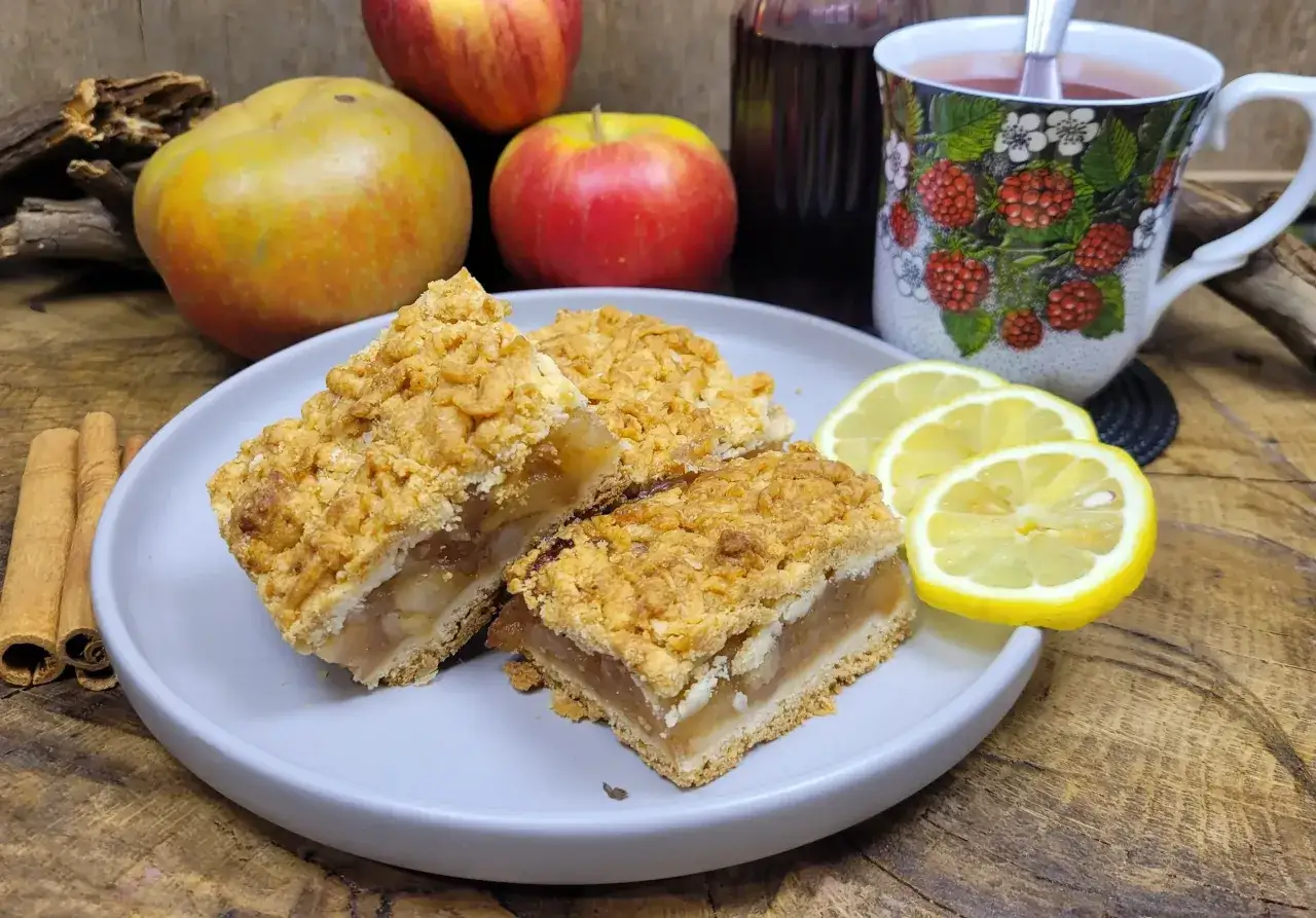 Kawałki pysznej szarlotki na talerzu, obok jabłka, cytryny i cynamonu. Idealne na prosty przepis na szarlotkę.
