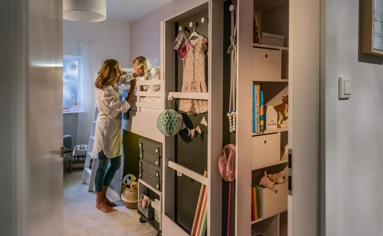 Mama i córka w małym pokoju dziecięcym w bloku. Na łóżku piętrowym, obok wiszą ubrania i ozdoby.