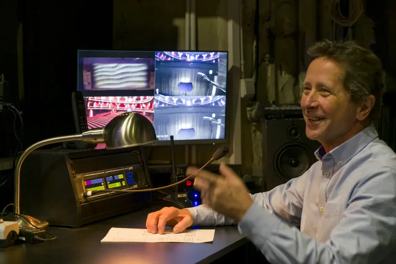 Uśmiechnięty mężczyzna przy konsoli dźwiękowej. Widać monitory z widokiem na scenę. Tak właśnie wygląda praca w teatrze.