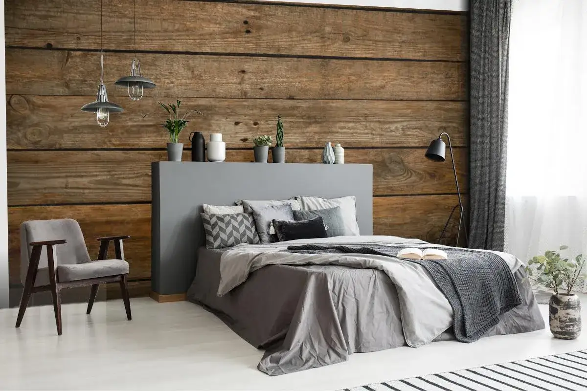 Sypialnia z tapetą stare deski na ścianie. Szare łóżko z poduszkami, fotel i lampy tworzą przytulną atmosferę.