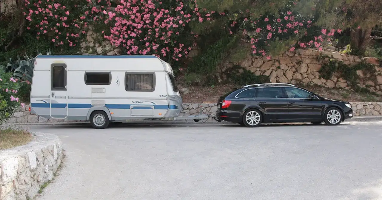 Czarny samoch&oacute;d ciągnie biały kamper kat C. W tle kwitnące krzewy i kamienny murek.